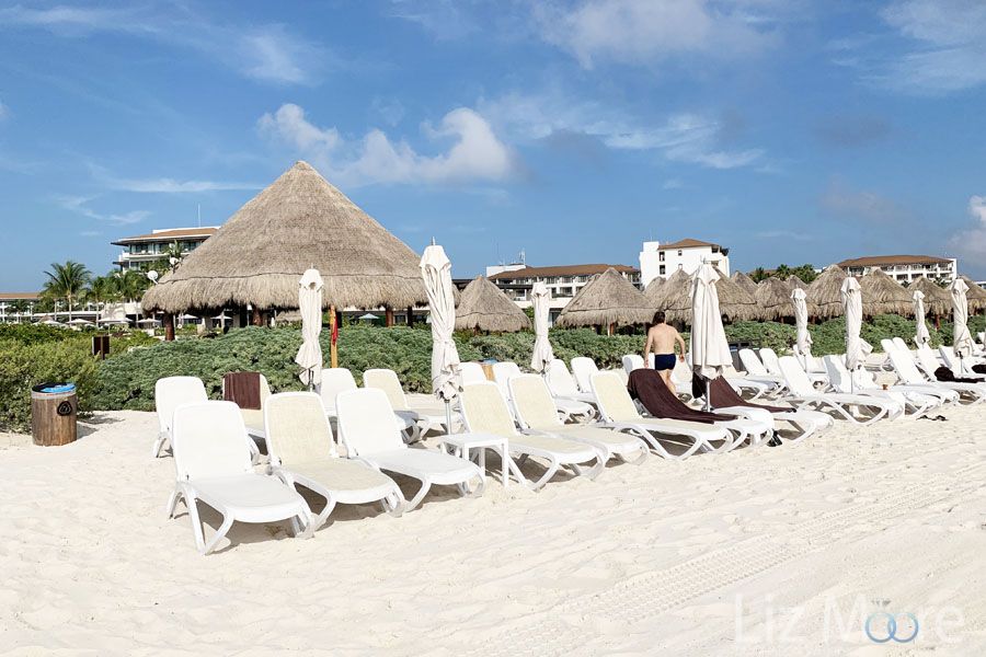 Many beach lounge chairs set up on the white sand beach by the ocean with resort in the background